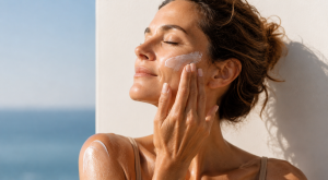 Femme en préménopause appliquant de la crème solaire sur son visage sous un soleil doux, avec une peau lumineuse et protégée en bord de mer.